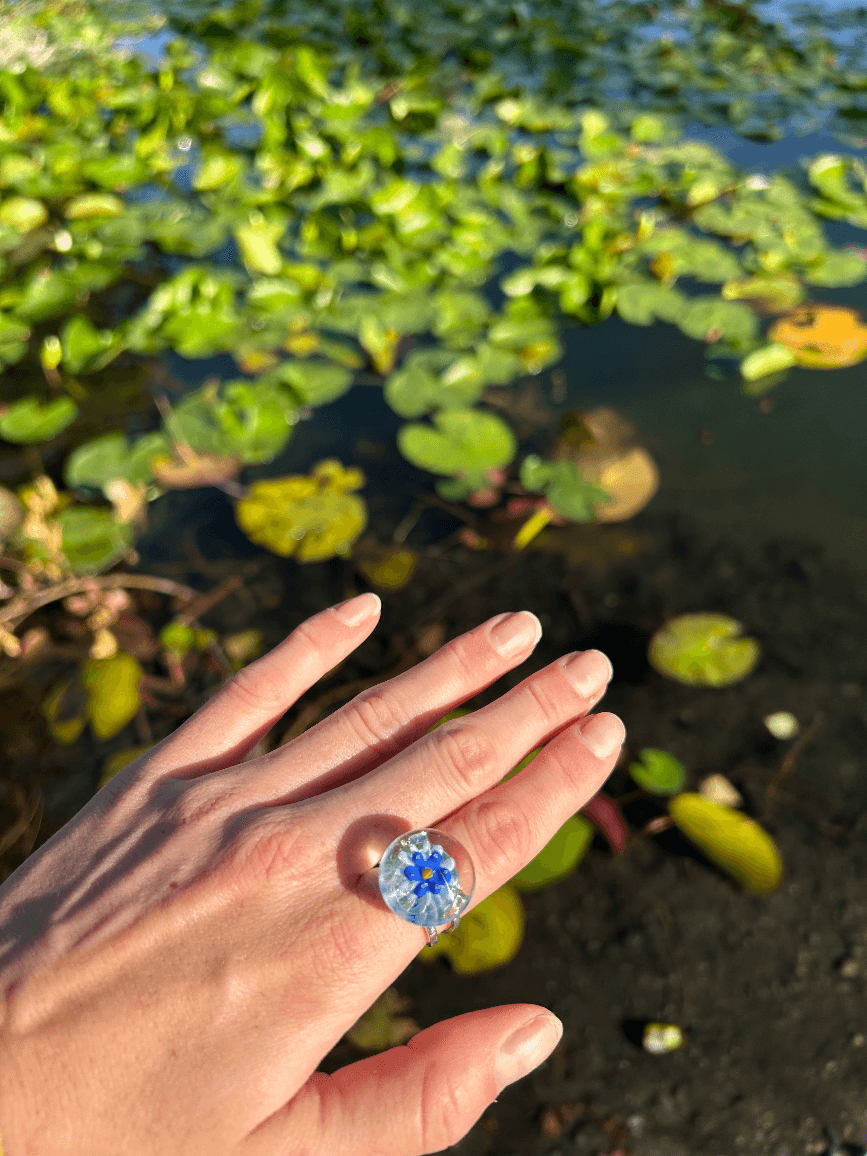Bague en verre portée à la main avec cabochon rond à fleur bleue et jaune, fond de nénuphars.