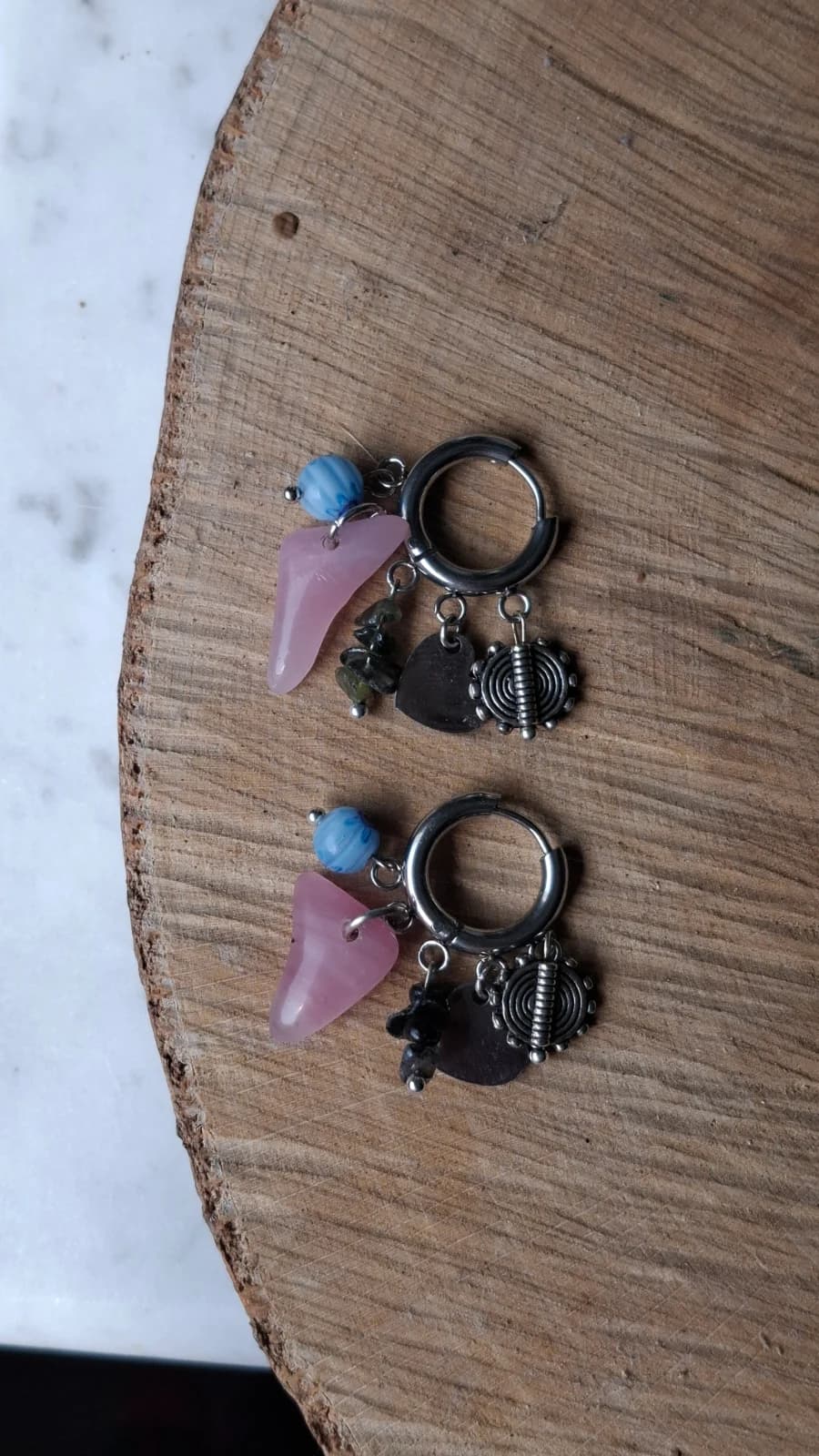 Paire de boucles d'oreilles en acier avec verre rose poli, perle bleue et pampilles sur un plateau en bois.