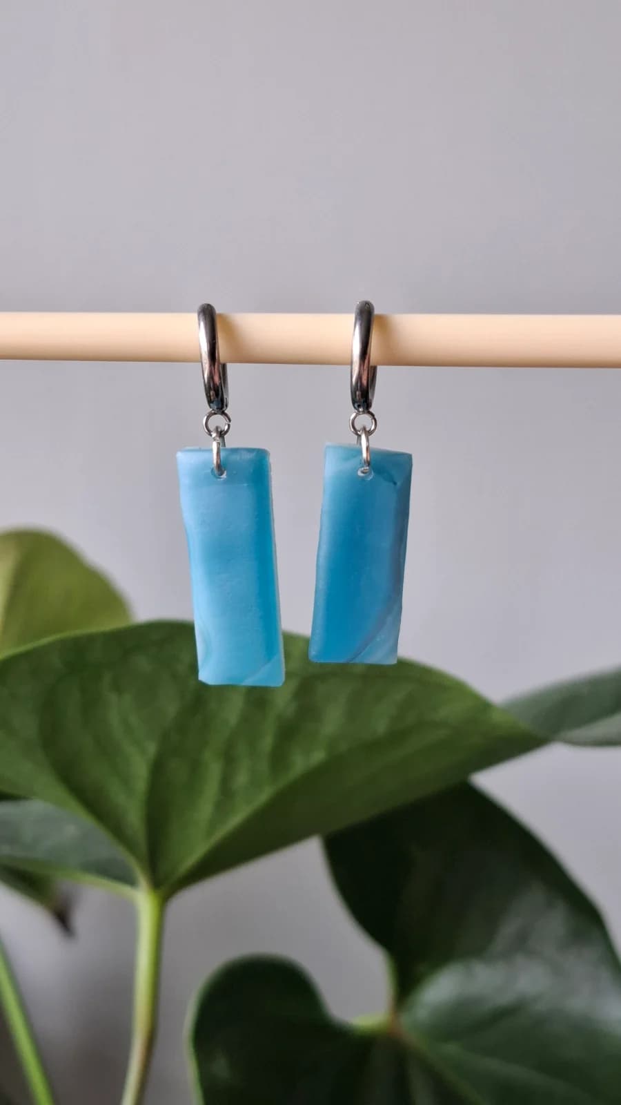 Boucles d'oreilles pendantes rectangulaires en verre bleu suspendues devant des feuilles vertes.