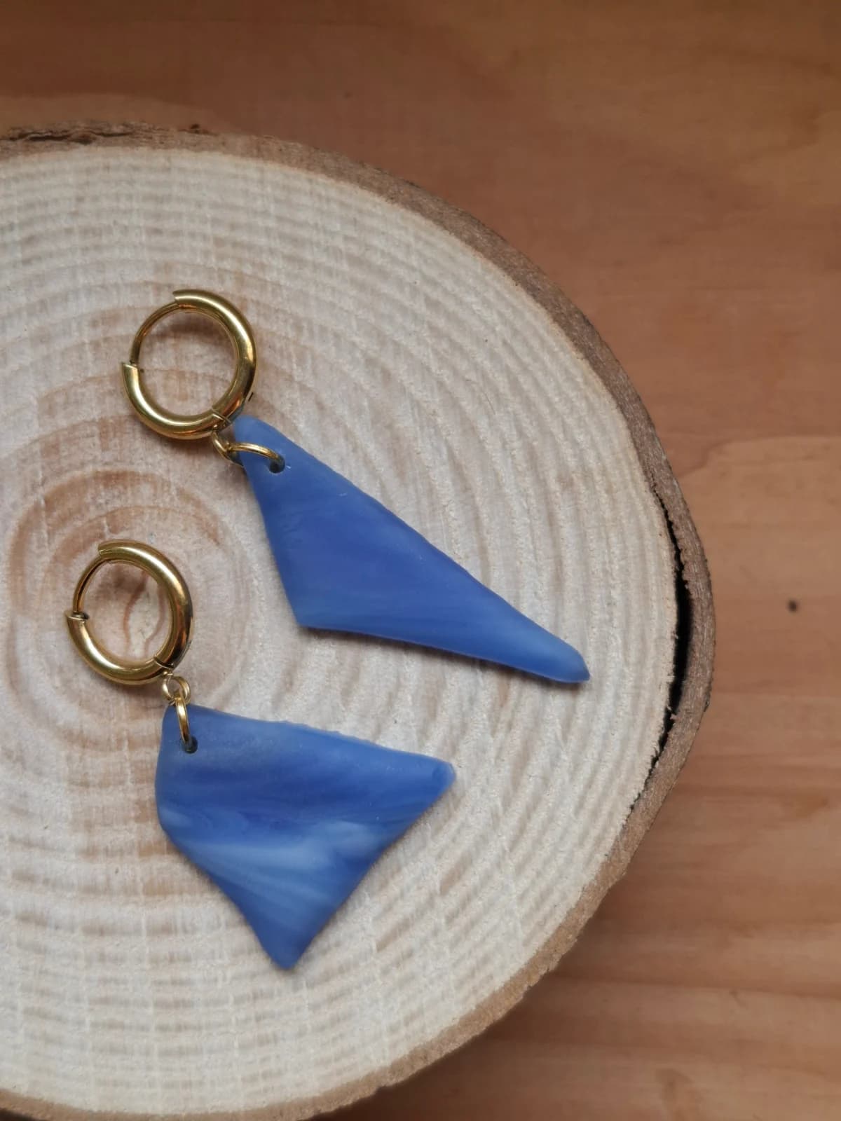 Boucles d'oreilles pendantes asymétriques en verre bleu marbré posées sur une tranche de bois.