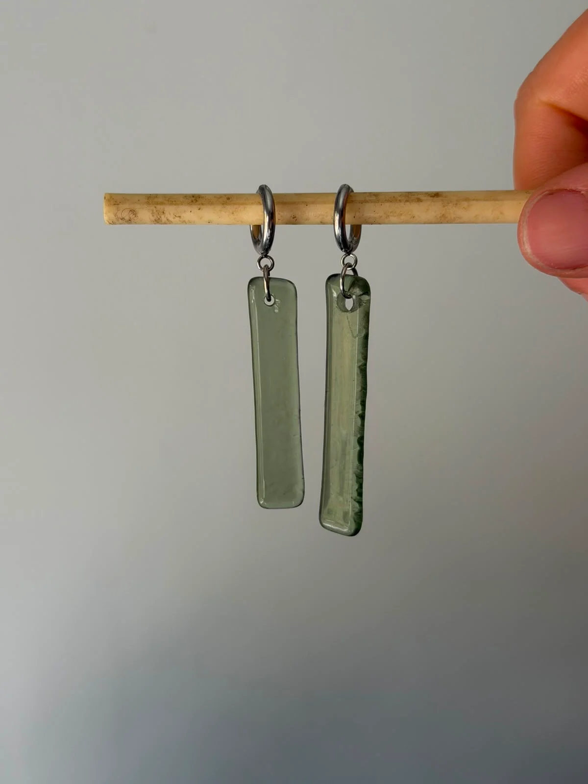 Boucles d'oreilles pendantes vertes présentées sur un support doré devant un voilage blanc.
