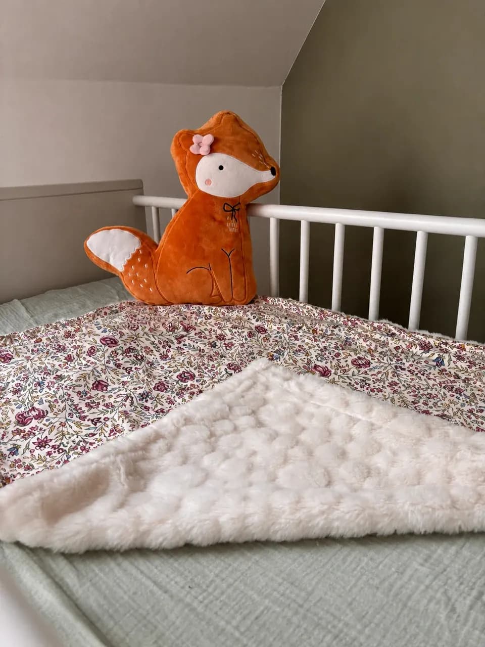 Couverture bébé à motif floral et polaire blanche texturée posée sur un lit avec un coussin renard.