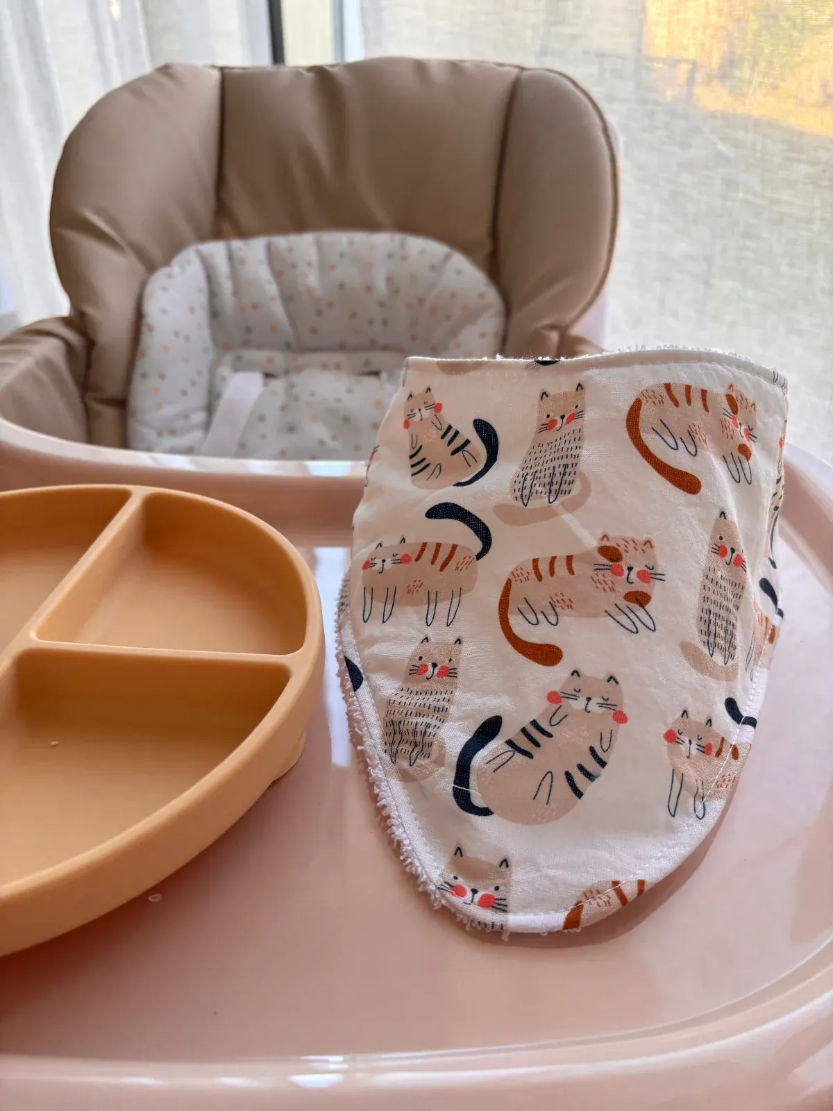 Bavoir bandana blanc à motifs de chats illustrés, posé sur le plateau d'une chaise haute pour bébé.