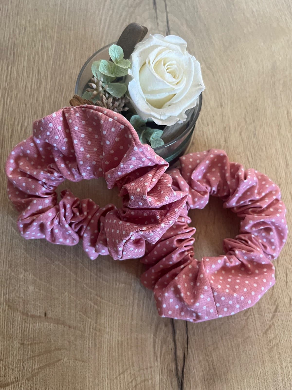 Deux chouchous froncés roses à pois blancs posés sur une table en bois près d'une rose blanche.