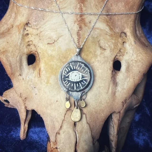 Gros plan sur le pendentif talisman orné d'un œil gravé et de trois gouttes dorées suspendues.
