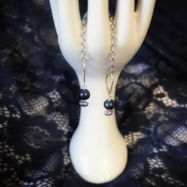 Boucles d'oreilles pendantes aux perles sombres présentées sur une main décorative blanche sur fond noir.