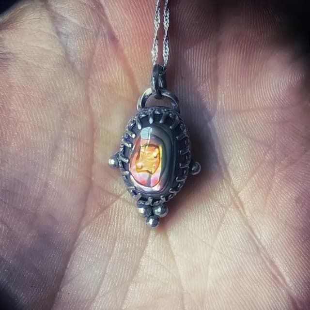 Pendentif en argent patiné serti d'une abalone aux reflets dorés et orangés, posé au creux d'une main.