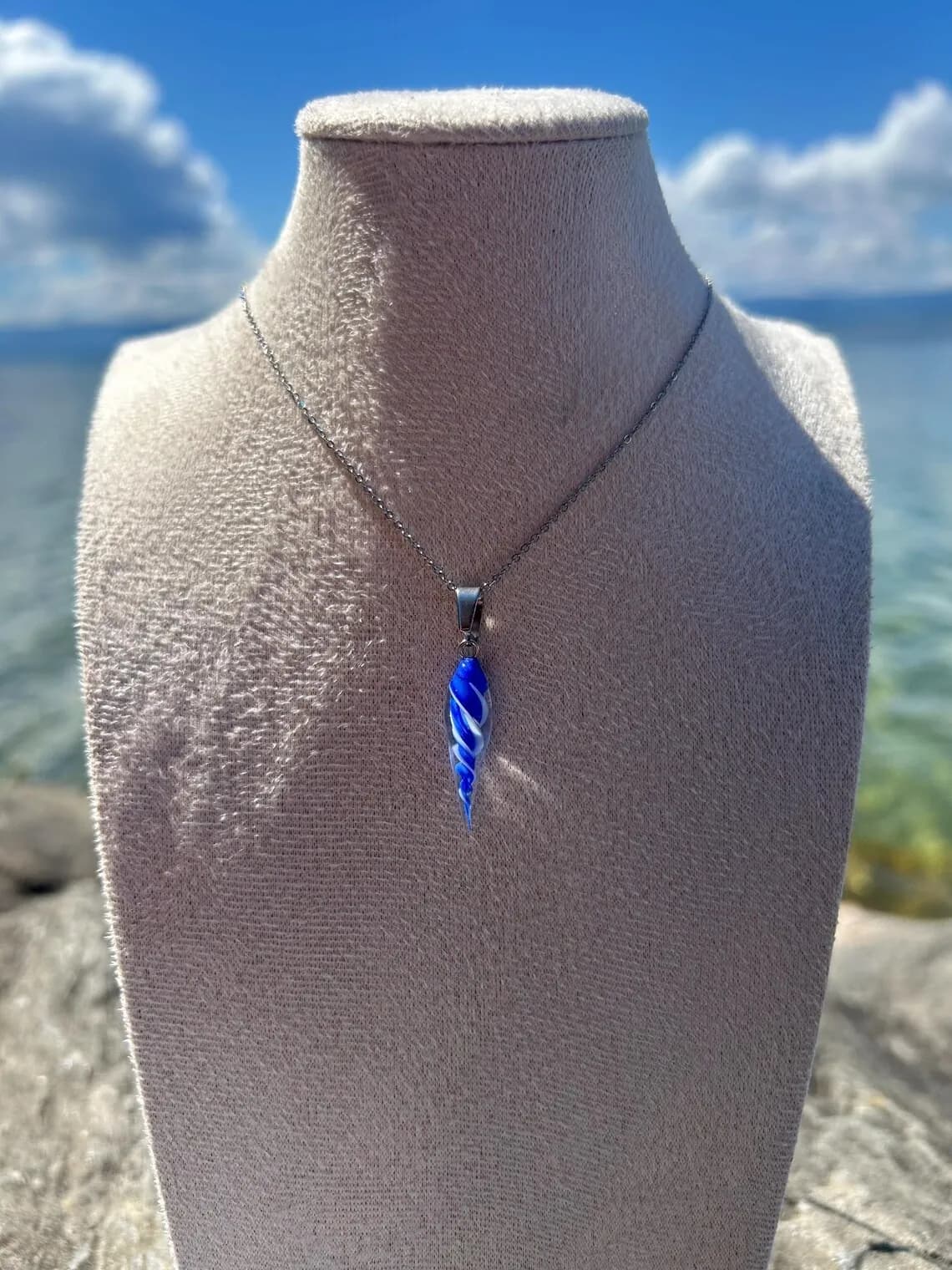Collier avec pendentif coquillage bleu et blanc en verre filé sur un buste d'exposition face à l'eau